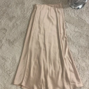 Champagnefärgad långkjol från Vila - Säljer en elegant långkjol från Vila i en mjuk champagnefärgad ton. Kjolen har en slät, glansig yta och är tillverkad i ett satinliknande material som ger en lyxig känsla. Passformen är normal och kjolen har en enkel, stilren design utan mönster eller extra detaljer. Använt en gång 