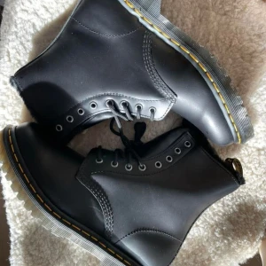 Dr. Martens boots med varmt foder - Säljer ett par mörkgrå Dr. Martens boots (ser svarta ut) med klassisk gul söm runt sulan och snörning framtill. Skorna har rund tå, platt sula och är fodrade med svart päls för extra värme. Tillverkade i skinn och har robust yttersula som ger bra grepp. Perfekta för kalla dagar. Storlek 41. Helt nya. Skickas i orginalkartong. Kostar 2200kr nya.
