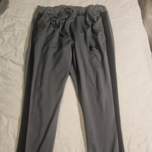 Grå träningsbyxor från Under Armour  - Grå träningsbyxor från Under Armour i storlek XL men passar som L. Byxorna har elastisk midja med snörning, raka ben och svart logga på låret. Fast pris 190 kr.