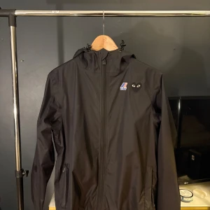 Svart vindjacka Comme des Garçons x K-Way - Svart vindjacka från Comme des Garçons Play i samarbete med K-Way. Jackan har huva med dragsko, hel dragkedja framtill och två sidofickor. Ikoniskt hjärta med ögon på bröstet och K-Way-logga. Tillverkad i lätt polyester, perfekt för blåsiga dagar. Köpt på NK