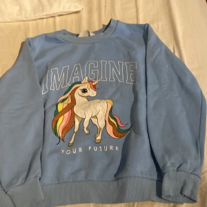 Blå tröja med enhörning från H&M - Superfin ljusblå sweatshirt från H&M med stort tryck av en enhörning och texten 'IMAGINE YOUR FUTURE' på bröstet. Tröjan har långa ärmar, ribbade muddar och är gjord i mjuk bomull. Perfekt för dig som älskar färgglada detaljer och magiska motiv!