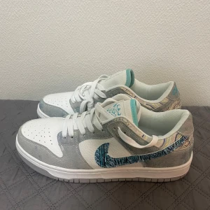 Nike Dunk Low i vitt och grått med mönster - Nike Dunk Low sneakers i vitt och ljusgrått skinn med texturerad yta. Swoosh-loggan har ett blått och svart mönster som sticker ut, och detaljer i turkos på plösen. Klassisk låg modell med snörning och perforerad tå för extra stil.
