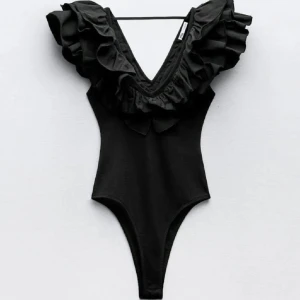 Svart body med volang och v-ringning från Zara  - Supersnygg svart bikini med djup v-ringning och stora volangdetaljer runt axlar och hals. Ger en trendig och unik look på stranden eller vid poolen. Perfekt för dig som vill sticka ut och känna dig extra cool i sommar. 