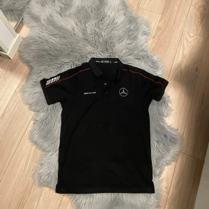 Svart Mercedes AMG Petronas pikétröja - Svart pikétröja från Mercedes AMG Petronas Motorsport med broderad logga på bröstet och vita detaljer på ärmen. Röd linje längs axlarna och klassisk krage med knappar. Perfekt för dig som gillar motorsport och snygg streetstyle.