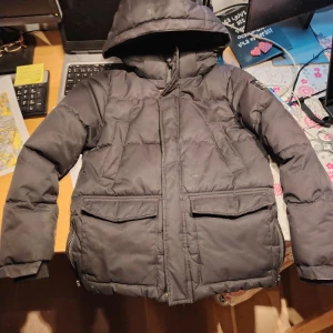 McKinley Svart Vinterjacka med Luva 140 - Svart pufferjacka från McKinley med huva och stora fickor framtill. Jackan har dragkedja, ribbade muddar och patch med logga på ärmen. Perfekt för kalla dagar och outdoor vibes. Jackan är nyskick förutom en fläck ovanför höger ficka. Se bilder. 