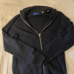 Svart stickad kofta från Jack & Jones - Svart stickad kofta från Jack & Jones med grov struktur och hel dragkedja framtill. Tröjan har ribbade muddar och krage, perfekt för lager-på-lager. Enkel och stilren design som funkar till det mesta.