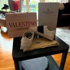 Valentino Garavani Open sneakers vit/marin - Snygga Valentino Garavani Open sneakers i vitt skinn med bred marinblå rem över vristen. Klassisk rund tå, vita snören och ikoniska nitar på hälen. Kommer med originalkartong och dustbag. Perfekta för dig som vill ha en clean men lyxig look.