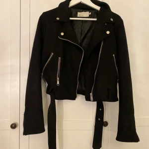 Svart bikerjacka från NLY Trend - Snygg svart bikerjacka från NLY Trend med croppad passform, silvriga dragkedjor och bälte i midjan. Jackan har långa ärmar med dragkedjor vid ärmsluten och coola detaljer i metall. Perfekt för dig som vill ha en edgy look.