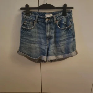 Blå högmidjade jeansshorts H&M - Säljer ett par klassiska blå jeansshorts från H&M i storlek 36. Modellen är högmidjad med uppvikta ben och fem fickor. Shortsen har knapp och dragkedja framtill samt bälteshällor. Perfekta för varma dagar och har en chill, tidlös vibe.