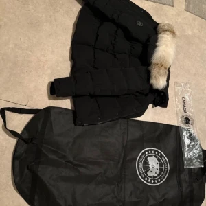 Svart dunjacka från Canada Goose - Säljer en svart dunjacka från Canada Goose med fluffig päls på huvan. Jackan har klassisk pufferstil, dragkedja och knappar framtill samt logga på ärmen. Perfekt för kalla vinterdagar och riktigt snygg till streetwear. Dragkedjan är dock lite sönder men man kan dra upp den enkelt såfort man försöker.