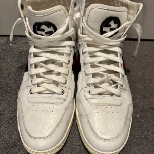 Vita high top sneakers från Gucci - Säljer ett par vita high top sneakers från Gucci i skinn med klassiska gröna och röda ränder på sidan samt svart Gucci-logga på plösen. Skorna har rund tå, platt sula och kommer med vita skosnören och originalpåse. Intyg på att varan är äkta finns https://backend.authentifier.com/originals/M-/68/ed/M-68ed71b952883.pdf?1760720467       