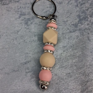 Nyckelring med pärlor i rosa och beige - Unik nyckelring med silikonpärlor i ljusrosa och beige, dekorerad med glittriga silverdetaljer. Pärlorna har både rund och geometrisk form, vilket ger en cool och modern vibe. Perfekt accessoar för att piffa upp din väska eller nyckelknippa.