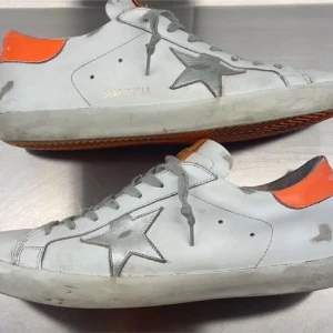 Golden Goose vita sneakers med orange detalj - Golden Goose sneakers i vitt med silverfärgad stjärna på sidan och orange hälkappa. Klassisk låg modell med grå snörning och platt sula. Snygg kontrast mellan det vita och den starka orangea detaljen, perfekt för dig som gillar streetstyle.
