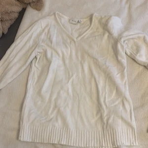Vit v-ringad stickad tröja från Vila - Vit stickad tröja från Vila med v-ringning och långa ärmar. Tröjan har ribbade muddar vid ärmslut och nederkant, vilket ger en snygg och enkel look. Perfekt att matcha med jeans eller kjol för en clean stil. Dock är den lite nopprig, vilket kan ses i sista bilden.
