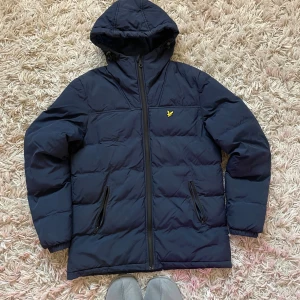 Marinblå pufferjacka från Lyle & Scott - Säljer en marinblå pufferjacka från Lyle & Scott med huva och dragkedja. Jackan har två fickor med dragkedja och den klassiska gula loggan på bröstet. Perfekt för kyliga dagar och riktigt snygg passform.