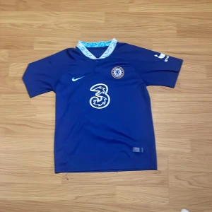 Chelsea Kanté #7 matchtröja Nike - Chelsea FC fotbollströja med Kanté #7 på ryggen, tillverkad av Nike. Tröjan är marinblå med vit och ljusblå detaljer på kragen, klubbmärke på bröstet och sponsorloggor. Materialet är lätt och andas, perfekt för träning eller match.