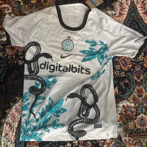 Inter Milan fotbollströja  - Snygg vit Inter Milan fotbollströja från Nike med svart ormmotiv och turkosa blad. Tröjan har svart krage och ärmar, digitalbits-logga på bröstet och klubbmärke. Tillverkad i lätt och ventilerande material, perfekt för match eller träning.