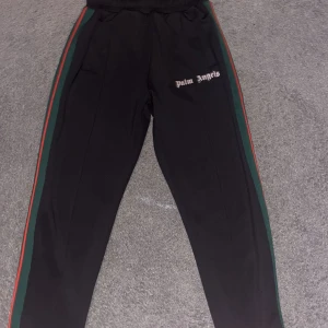 Palm Angels svarta byxor med ränder - Svarta byxor från Palm Angels med gröna och röda ränder längs sidorna. Byxorna har elastisk midja och broderad Palm Angels-logga på framsidan. Materialet är glansigt och känns sportigt, perfekt för en streetwear-look.