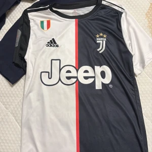 Juventus Ronaldo 7 matchtröja Adidas - Juventus fotbollströja med Ronaldo 7 på ryggen, Adidas-logga och Jeep som sponsor. Tröjan är svart och vit med en röd linje i mitten, korta ärmar och broderat Juventus-emblem med tre guldstjärnor. Materialet är lätt och andas, perfekt för match eller träning.