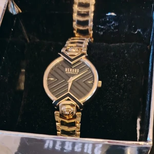 Versace guldig klocka med svart urtavla - Snygg klocka från Versus Versace för 2200kr med guldig länk och svart urtavla med randigt mönster. Klockan har unika detaljer som lejonhuvud på länken och geometriska former runt urtavlan. Perfekt accessoar för dig som gillar lyxig och modern stil.