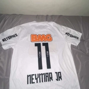 Vit Santos Neymar Jr fotbollströja Nike - Säljer en vit Santos fotbollströja från Nike med Neymar Jr och nummer 11 på ryggen. Tröjan har korta ärmar, Dri-Fit-material och tryckta sponsorer som BMG och Netshoes. Perfekt för dig som gillar brasiliansk fotboll och klassiska tröjor.