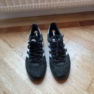 Adidas Samba sneakers i svart mocka - Säljer ett par klassiska Adidas Samba sneakers i svart mocka med vita ränder på sidorna och bruna gummisulor. Skorna har rund tå, snörning och låg profil. Perfekta för dig som gillar retrostil och sportig vibe.