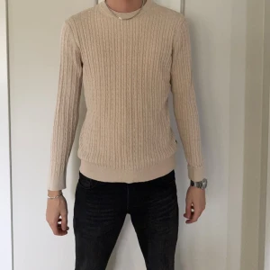 Beige stickad tröja från Jack & Jones - Säljer en beige stickad tröja från Jack & Jones Premium i storlek L. Tröjan har ett klassiskt kabelstickat mönster, rund halsringning och ribbade muddar vid ärmslut och nederkant. Perfekt för lager-på-lager under hösten och vintern.