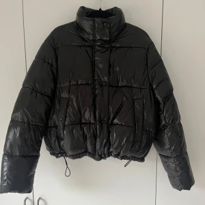 Svart pufferjacka med hög krage - Snygg svart pufferjacka med glansig finish och hög krage. Jackan har dragsko i nederkant, tryckknappar framtill och två fickor. Perfekt för kalla dagar när du vill hålla stilen och värmen.