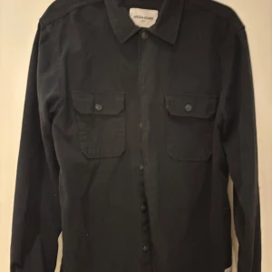 Svart overshirt från Jack & Jones - Svart overshirt från Jack & Jones med klassisk krage och två stora bröstfickor med knappstängning. Skjortan har långa ärmar och knappar framtill. 