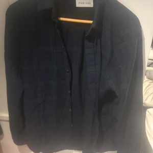 Mörkblå rutig skjorta från Pier One - Säljer en mörkblå rutig skjorta från Pier One med klassisk krage och knappar framtill. Skjortan har lång ärm och en loose passform som sitter chill på kroppen. Perfekt för dig som gillar en avslappnad stil med lite edge. Materialet är mjukt och känns skönt mot huden.