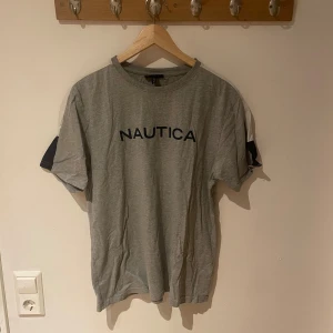 Grå t-shirt från Nautica - Snygg grå t-shirt från Nautica med klassisk rund hals och tryckt logga på bröstet. T-shirten har korta ärmar och är gjord i mjuk bomull, perfekt för en chill och stilren look. Passar dig som gillar enkel design med en touch av street.