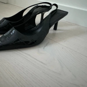 Svarta pumps med spetsig tå - Säljer ett par svarta pumps med spetsig tå och öppen häl. Skorna har en låg klack och ett blankt, krokodilpräglat material som ger en cool vibe. Perfekta för dig som gillar stilrena och trendiga skor med lite edge.