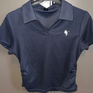 Mörkblå polotopp från SHEIN - Mörkblå polotopp från SHEIN med klassisk krage och korta ärmar. Toppen har en vit broderad detalj på bröstet och är gjord i ett mjukt, syntetiskt material. Perfekt för en clean och sportig look.