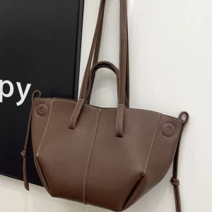 Brun axelväska i skinn - Snygg brun axelväska med stilren design och praktiska handtag. Väskan är tillverkad i skinn med synliga kontrastsömmar och har knyt-detaljer på sidorna. Perfekt storlek för att få plats med allt du behöver under dagen.