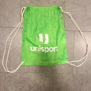 Grön gympåse från Unisport - Säljer en grön gympåse från Unisport med vitt tryck och snörning. Perfekt för att bära träningskläder eller skor. Väskan har en enkel och smidig design med vita snören som axelremmar.