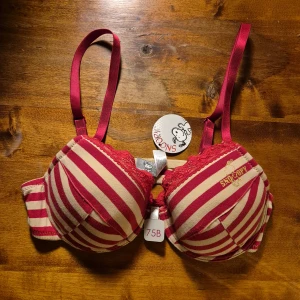 Randig röd och beige BH Snoopy 75B - Säljer en söt randig BH från Seppälä med Snoopy-tryck i guld på ena kupan. Toppen är röd och beige med spetsdetaljer upptill och justerbara axelband. Materialet är mjuk bomull med lite elastan för extra komfort. Perfekt för dig som gillar roliga detaljer!