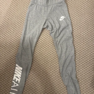 Grå Nike Air leggings - Grå leggings från Nike Air med vit logga på låret och längs benet. Tight passform och hög midja, perfekta för träning eller chill. Tillverkade i mjukt och stretchigt material för maximal komfort.
