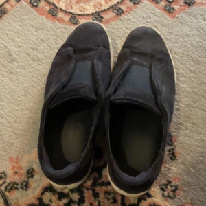 Axel arigato  - Svarta slip-on sneakers med vit sula, tillverkade i mocka och syntet. Skorna har en rund tå och platt sula, med elastiska paneler på ovansidan för enkel på- och avtagning. Perfekt för dig som gillar enkel och stilren design inte jätte bra skick men funkar att ha 