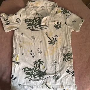 Vit kortärmad piké  från Warp - Vit piké t shirt  från Warp med coolt tryck av palmer, surfbrädor och graffiti-liknande text i grönt och gult. Skjortan har korta ärmar, klassisk krage, bröstficka och knappar framtill. Perfekt för en avslappnad och chill stil.