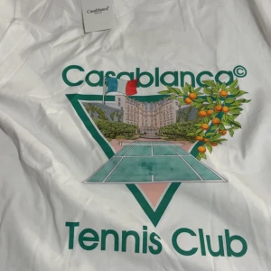 Vit Casablanca Tennis Club t-shirt - Vit t-shirt från Casablanca med stort färgglatt tryck på bröstet som visar en tenniscourt, apelsinträd och byggnad. Grön text 'Tennis Club' och 'Casablanca' framtill. Klassisk passform och rund hals, perfekt för en sportig look. Oanvänd 