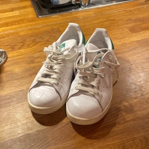 Adidas Stan Smith vita sneakers - Klassiska Adidas Stan Smith sneakers i vitt med gröna detaljer på hälen och tungan. Skorna har snörning, rund tå och platt sula. Tillverkade i skinn med perforerade sidor för ventilation. Se reva på bild 4. Använt skick och lite slitna.