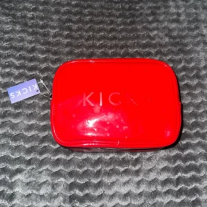 Röd necessär från Kicks med dragkedja - Snygg röd necessär från Kicks i blankt syntetmaterial. Väskan har en praktisk dragkedja och en stilren, rektangulär form. Perfekt för att organisera smink eller småprylar i väskan. Kicks-loggan syns tydligt på framsidan.