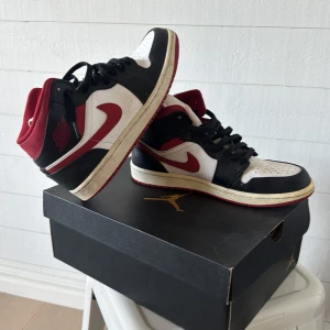 Nike Air Jordan 1 Mid Svart/Röd/Vit - Nike Air Jordan 1 Mid i klassisk svart, röd och vit färg. Skorna har svart snörning, röd swoosh och detaljer, samt vit tåbox och sidor. Tillverkade i skinn med textilfoder och ikonisk Air Jordan-logga på sidan. Perfekta för dig som gillar streetstyle och sneakerskultur.