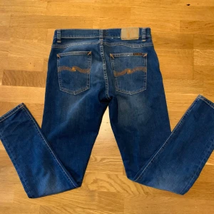 Blå Nudie Jeans, storlek 32/30 (Tight Terry) - Säljer ett par klassiska blå jeans från Nudie Jeans med snygga orange sömmar och ikonisk broderad bakficka. Modellen har normal passform och raka ben. Jeansen är tillverkade i bomull och har fem fickor samt läderpatch bak. Modell Tight Terry.