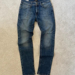 Jack & Jones Jeans Blå | W29 - Snygga blå skinny jeans från Jack & Jones, modell Liam. Jeansen har klassisk femficksdesign, skinny fit och en liten sliten detalj på ena benet. Perfekta för dig som gillar en tajt passform och stilren look.