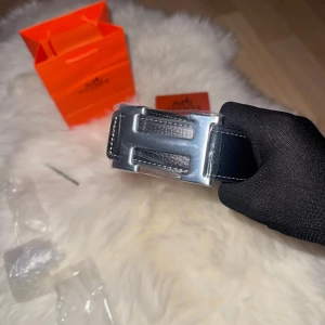 Svart H bälte med H-spänne - Säljer ett svart bälte från H med klassiskt H-spänne i silverfärgad metall. Bältet är gjort i skinn och har vita kontrastsömmar längs kanterna. Perfekt accessoar för att lyfta vilken outfit som helst. Inget i bilden ingår förutom bältet och håltagare 