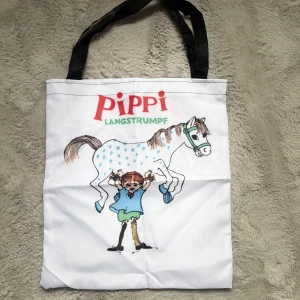 Pippi Långstrump tygkasse med tryck - Vit tygkasse med svart handtag och färgglatt Pippi Långstrump-motiv där Pippi lyfter sin häst. Trycket har blå, gröna, röda och bruna detaljer. Perfekt för dig som älskar klassiska barnboksfigurer och vill ha en unik och lekfull shoppingväska.