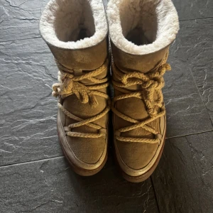 Beige Inuikii boots med snörning - Säljer ett par beige boots från Inuikii med grov snörning och mysigt foder. Skorna har en platt sula, rund tå och är tillverkade i mocka och skinn. Perfekta för kalla dagar med sin varma insida och snygga design.