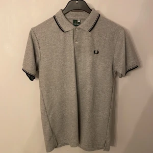 Grå pikétröja från Fred Perry M - Mer ljus grå i person Klassisk grå pikétröja från Fred Perry som stått i garderoben och dammat bort den är i bomull med svarta detaljer på krage och ärmslut. Tröjan har korta ärmar, två knappar vid halsen och den ikoniska Fred Perry-loggan broderad på bröstet. Perfekt för en clean och stilren look.