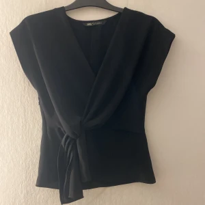 Svart omlottopp med knyt från Zara - Snygg svart omlottopp från Zara med v-ringning och knyt framtill. Toppen är ärmlös och har en dragkedja i sidan för enkel påklädning. Perfekt för dig som gillar stilrena och trendiga plagg. Har sytt ihop typ 1 cm på sidan, men det är inget som syns 